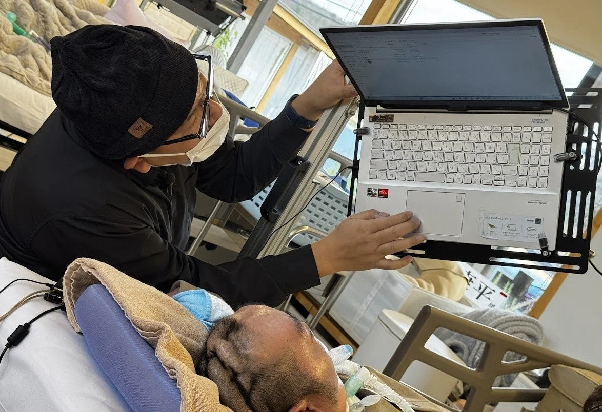 マスクとニット帽を着用した男性が、ベッドに横たわる患者の傍らでノートパソコンを操作しています。医療現場か自宅での介護中にPCで作業をしている様子です。
