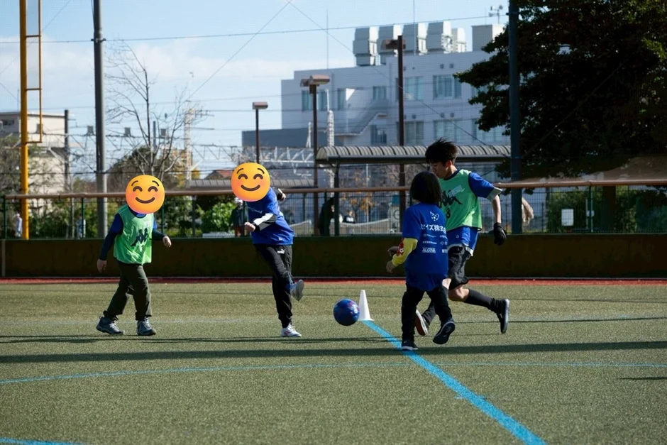 晴れた日に人工芝のグラウンドで子供たちがサッカーをしている様子。ボールとコーンを使って練習中か、楽しそうにプレーしている。