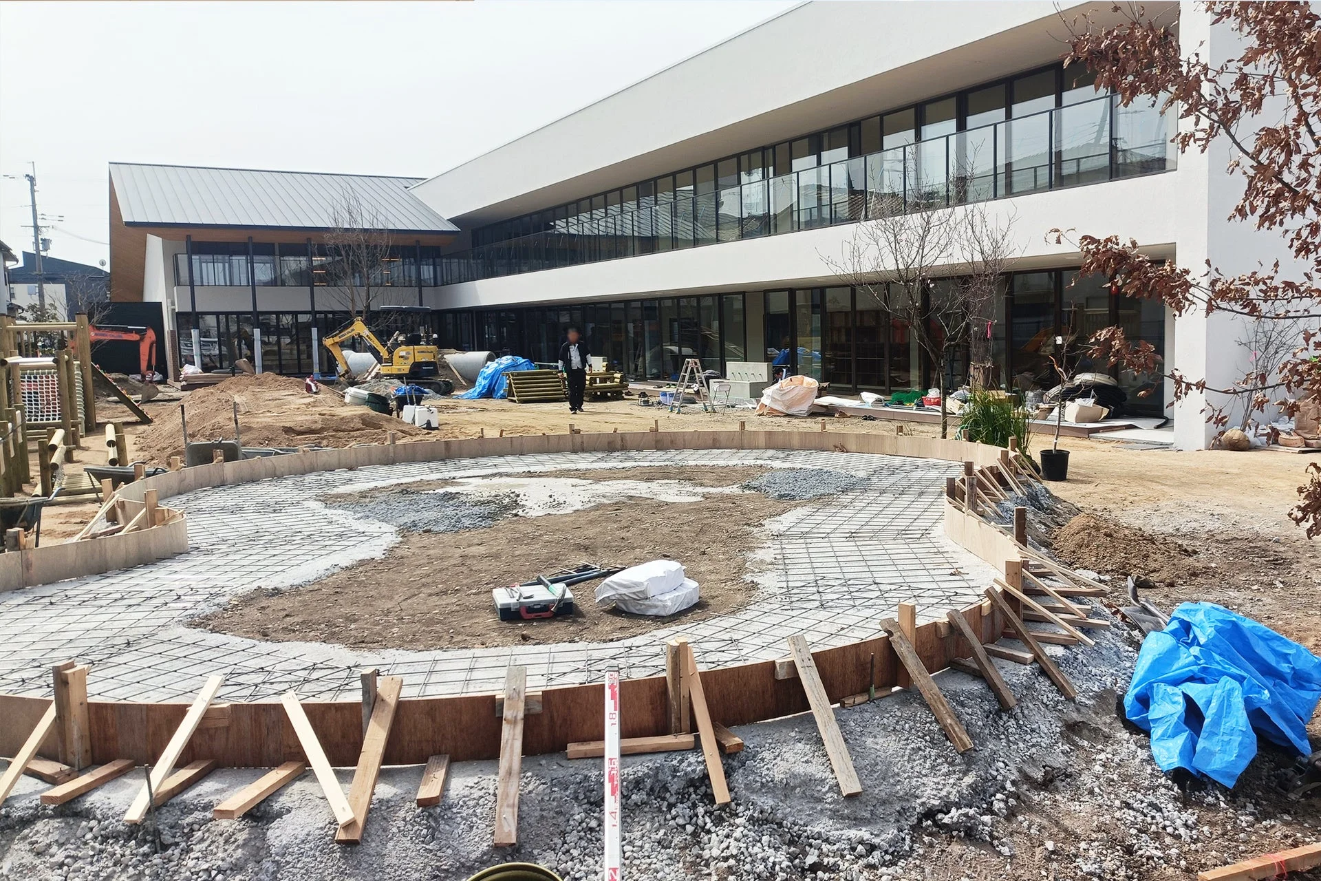 モダンなデザインの建物の前で、屋外エリアの建設工事が進められている様子。円形の基礎工事が行われ、ショベルカーや資材が置かれている。遊び場のような施設も見える。