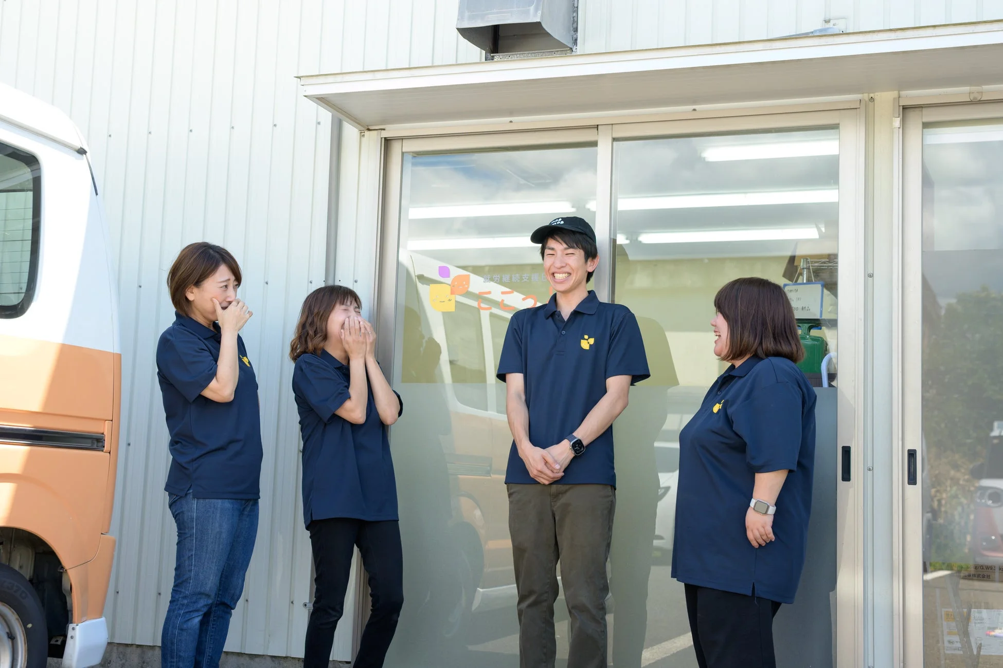 青いポロシャツを着た4人の人々が建物の前で笑顔で談笑