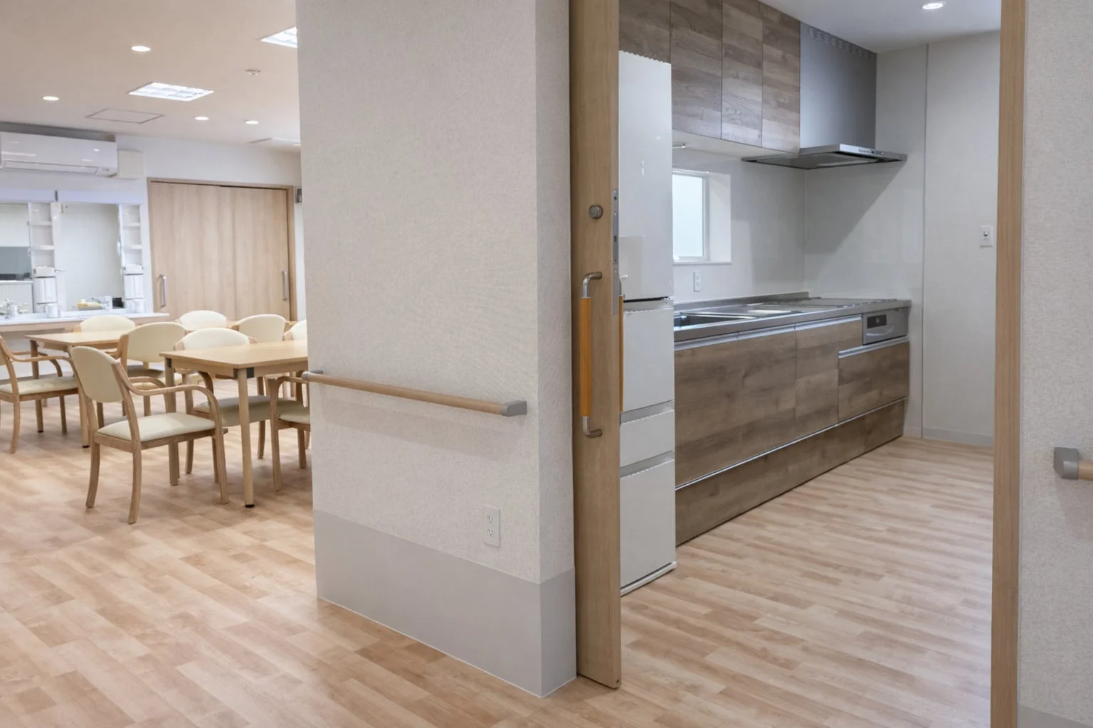 明るく清潔感のある共同スペースで、奥には木目調のダイニングテーブルと椅子が並び、手前にはモダンなキッチンが配置されている