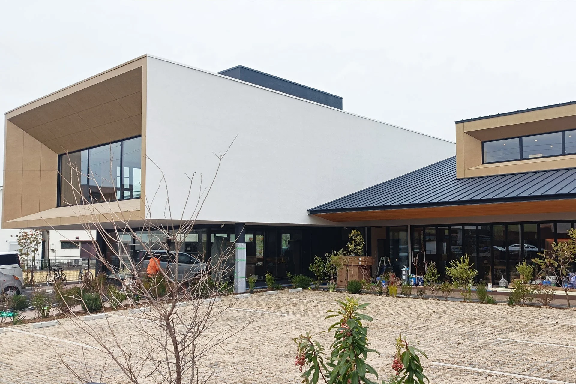 現代的で洗練されたデザインの商業施設と見られる建物の外観です。白い壁とベージュのアクセント、大きなガラス窓が特徴で、手前には舗装された駐車場と植栽があります。
