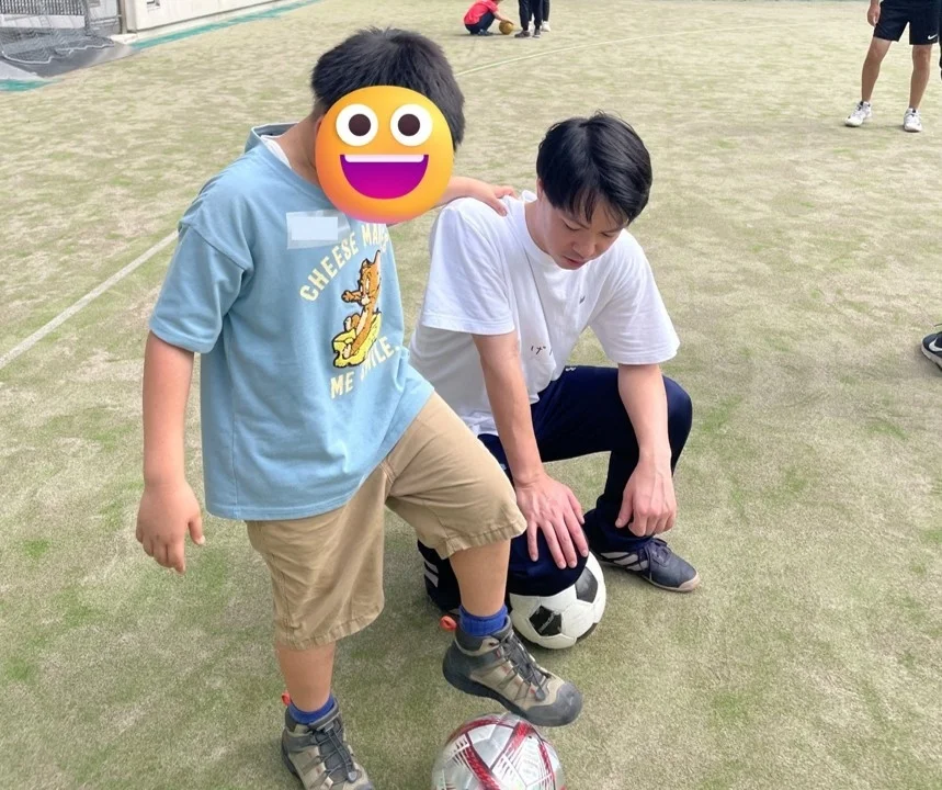 人工芝のグラウンドで、大人の男性が子供の肩に手を置き、サッカーボールを囲んで交流している様子です。スポーツ指導や練習の一場面のようです。