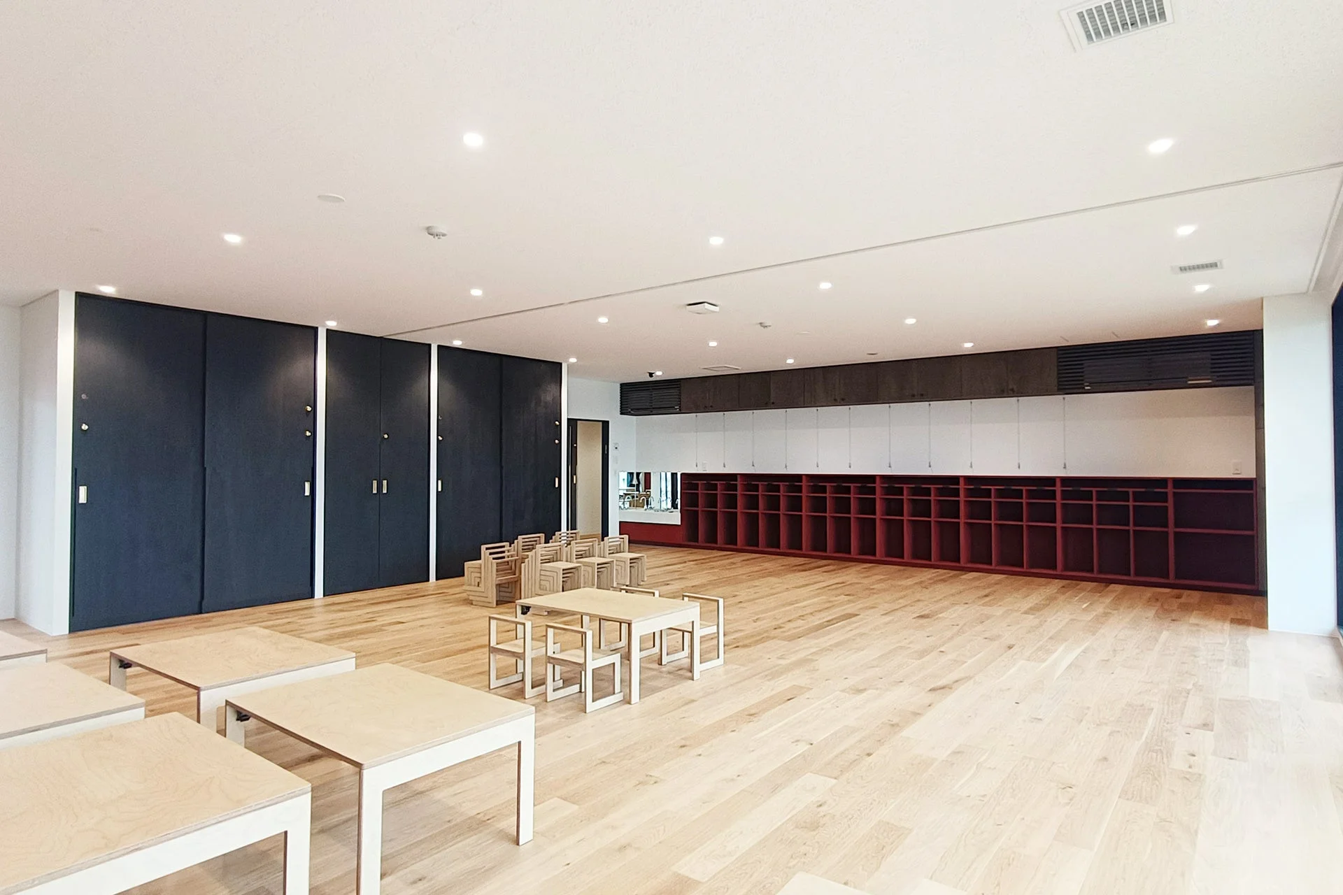 広々とした明るい室内空間で、木製の床にシンプルなテーブルと椅子が並んでいます。壁際にはロッカーや赤い収納棚があり、多目的室や教室として利用されている清潔感のあるモダンな空間です。