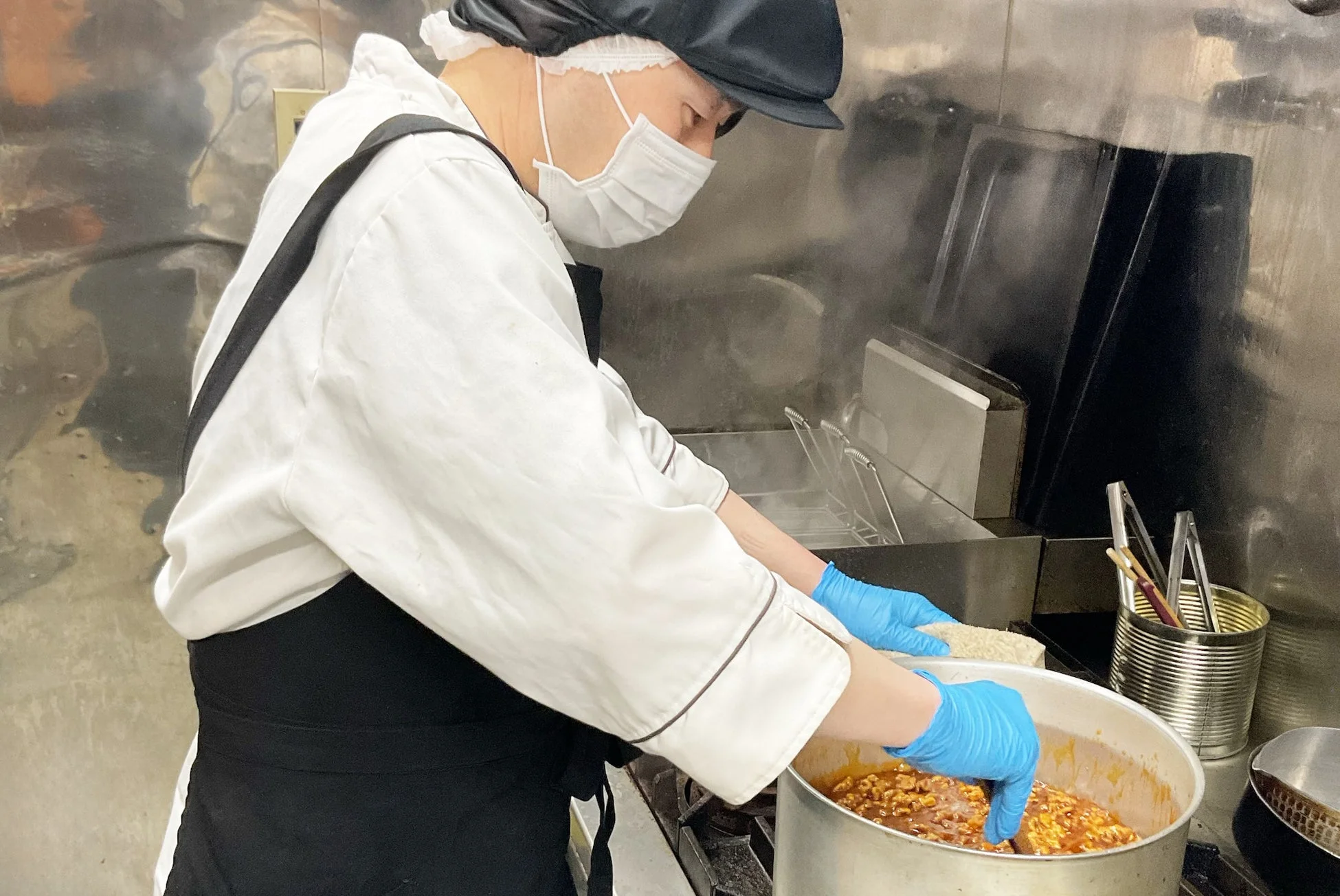 衛生的な服装の料理人が、マスクと手袋を着用し、厨房で大きな鍋に入った料理をかき混ぜています。