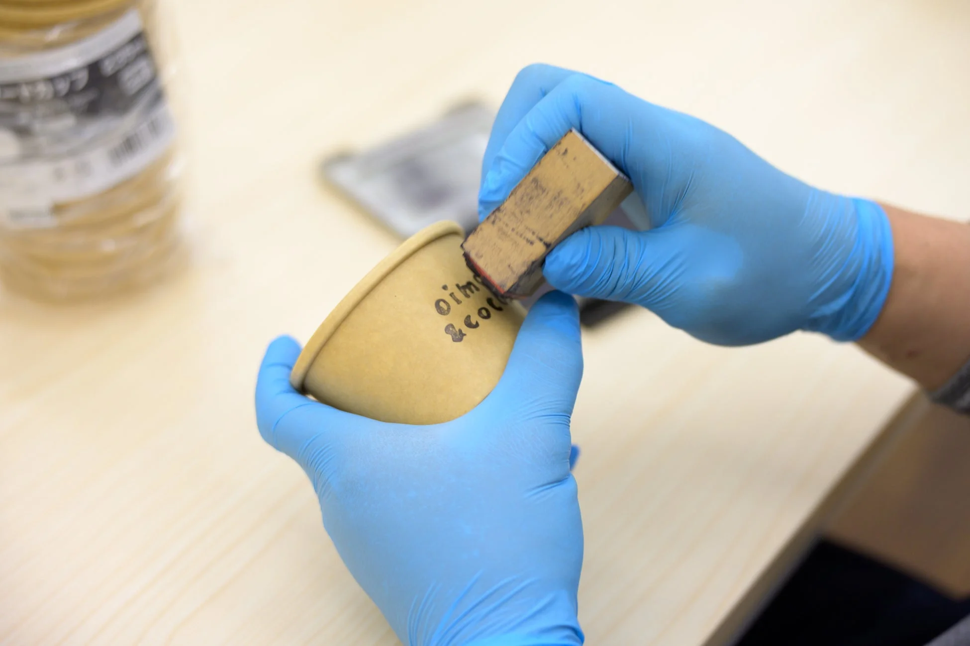 青い手袋をはめた手が、紙コップにロゴをスタンプで押している様子