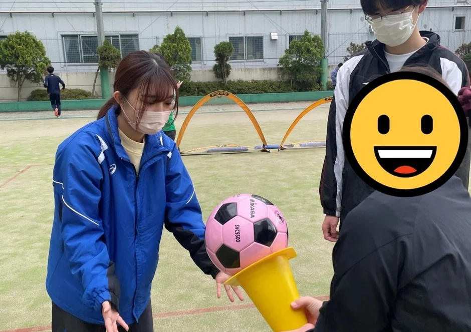 緑のグラウンドで、数人がスポーツ活動をしている様子です。青いジャケットを着た女性がピンクのサッカーボールに手を伸ばしており、近くにはマスクを着用した男性が立っています。背景には小さなサッカーゴールが見え、和やかな雰囲気の中でレクリエーションが行われているようです。