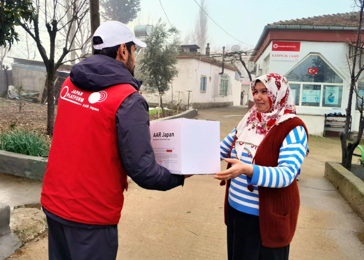 JAPAN PLATFORMとAAR Japanのロゴが入ったベストを着た男性が、ヒジャブを着用した女性に白い箱を手渡しています