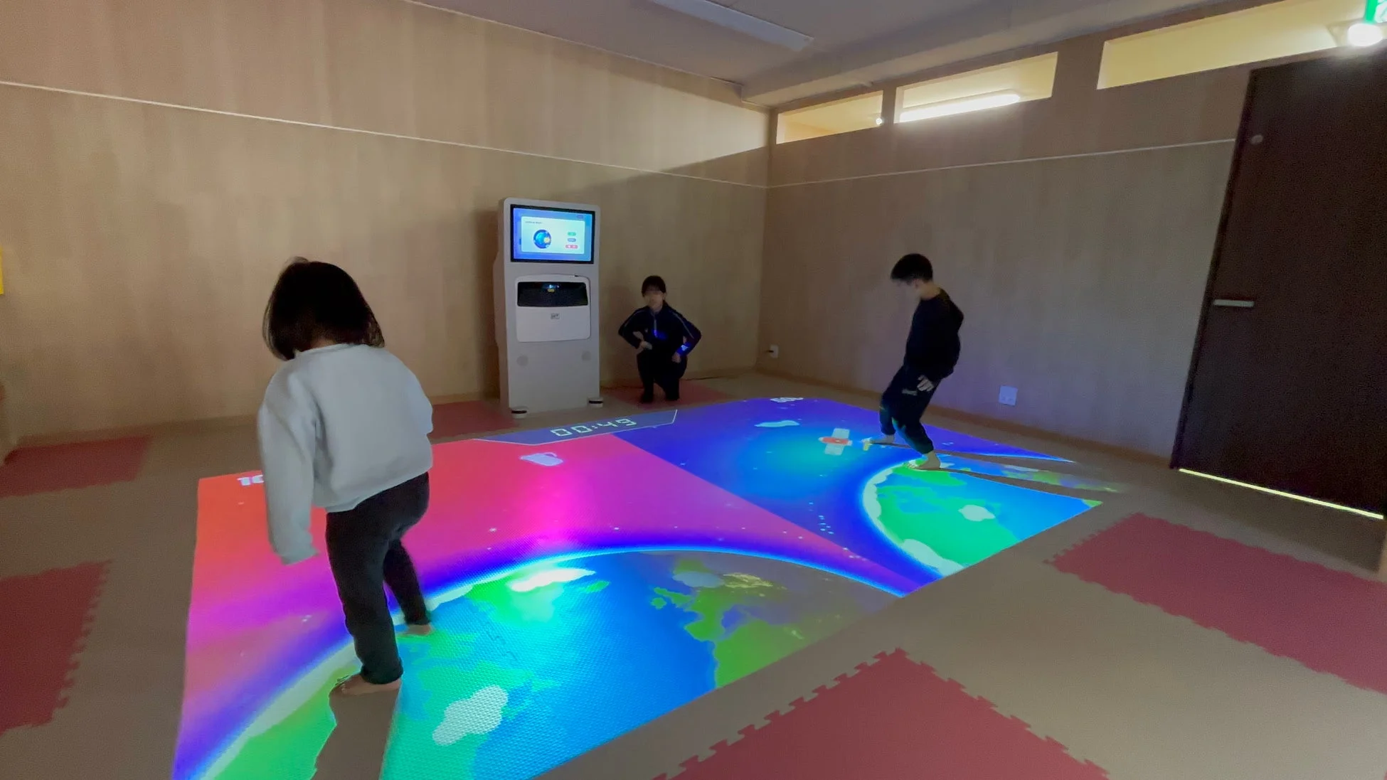子供たちが室内でインタラクティブな床面プロジェクションゲームを楽しんでいます。地球や宇宙をテーマにしたカラフルな映像が床に映し出され、子供たちはその上を動き回って遊んでいます。