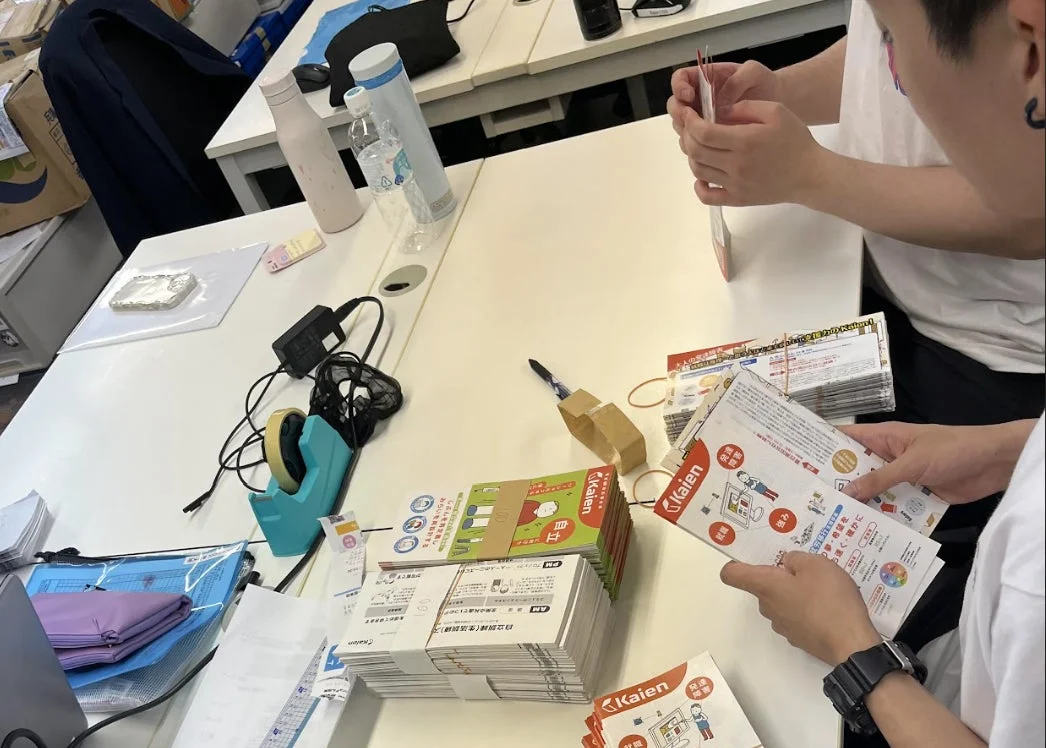 オフィスで二人が机に積まれた大量のパンフレットを仕分けたり準備したりしている様子