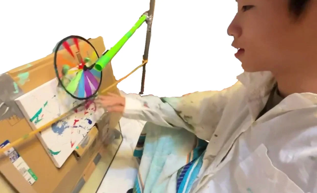 カラフルな風車と手作りの装置を使い、段ボール上のキャンバスに絵の具を飛ばしてアート作品を制作しているPOTEさん