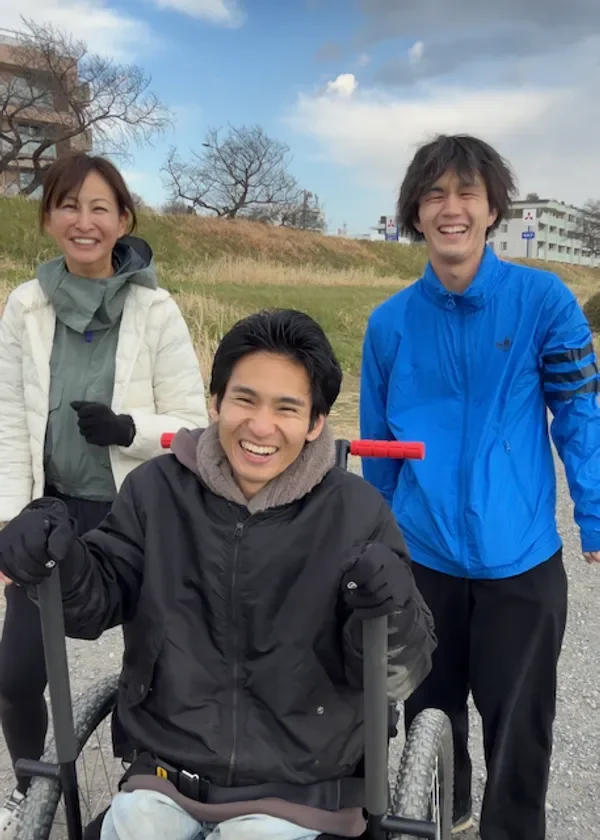 青空の下、屋外で3人の人物が笑顔で写っています。中央の男性は車椅子に乗っており、他の2人と共に楽しそうに散歩している様子が伺えます。