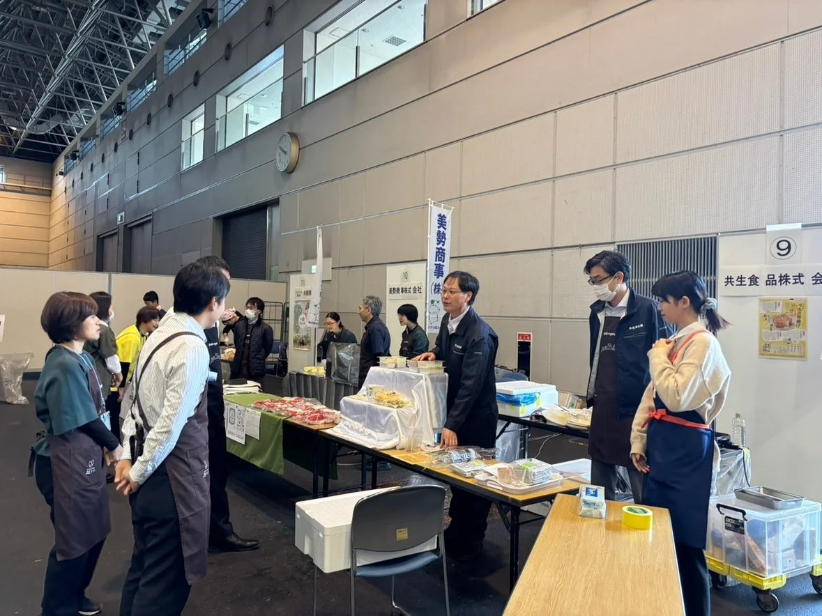 屋内会場で開催されている食品関連の展示会の様子。美勢商事や共生食品などの企業ブースが並び、スタッフが来場者と交流している。テーブルには肉や加工食品が展示され、活気ある商談が行われている。