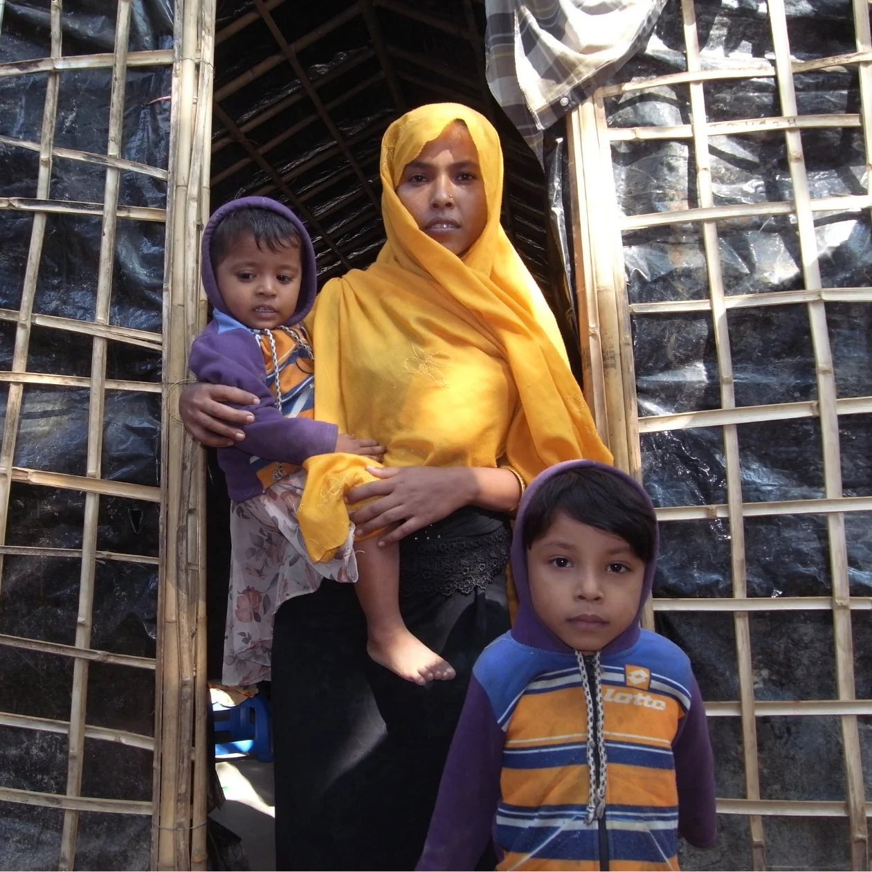 難民キャンプのような仮設住宅の前で、黄色いヒジャブを巻いた女性が幼い子供を抱き、もう一人の子供が隣に立つ家族のポートレート