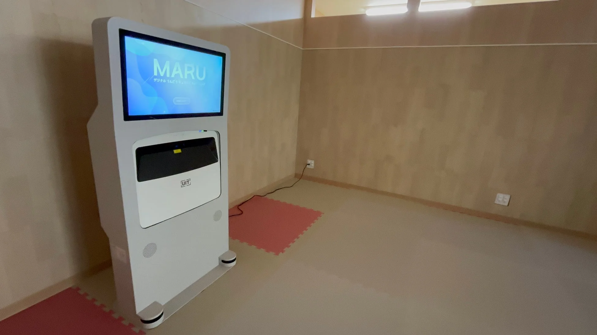 「MARU デジタルうんどうりょく」と表示されたデジタルサイネージが置かれたシンプルな室内。運動や健康に関連するサービスの端末と見られる。