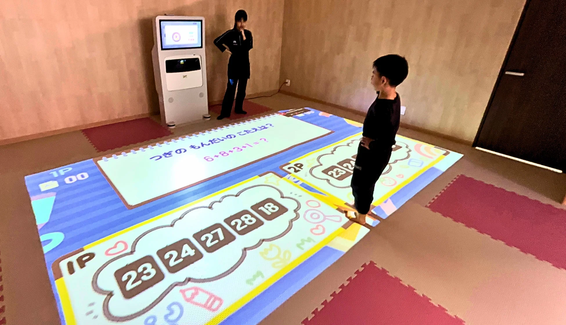 子供が床に投影されたインタラクティブな算数ゲームで遊んでいる様子です。女性がコンソール近くに立ち、見守っています。計算問題と選択肢が表示され、楽しみながら学べる空間が描かれています。