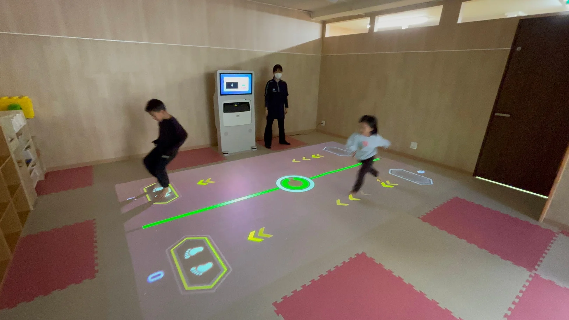 屋内で子供たちがインタラクティブな床プロジェクションゲームを楽しむ様子。足跡や矢印の指示に従い、体を動かすデジタルアトラクションで、大人が見守っています。
