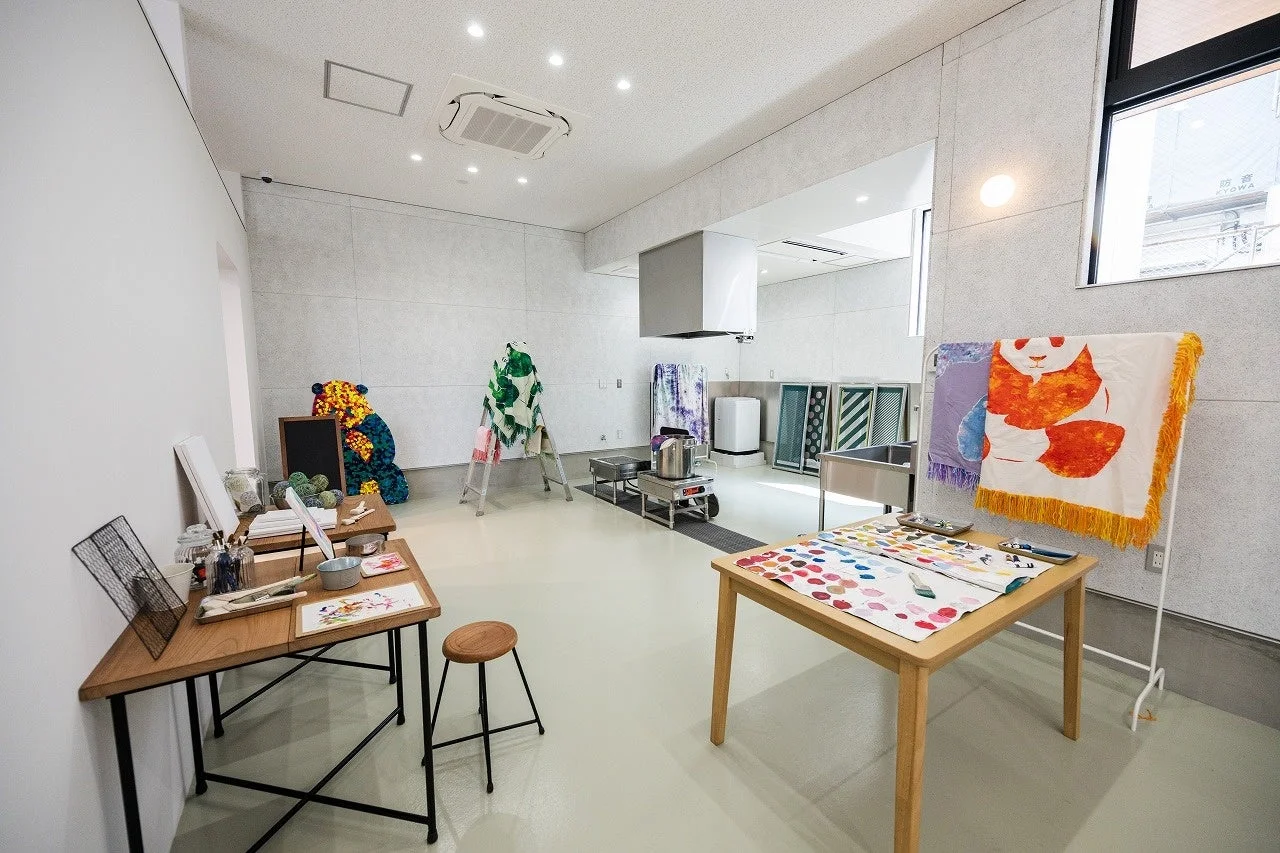広々とした明るいアートスタジオで、絵の具や筆が置かれた作業台、染物用のシンクや調理器具が見られます。カラフルなテキスタイルやパンダ柄のタオルが飾られ、創造的な活動が行われる工房の様子を捉えています。
