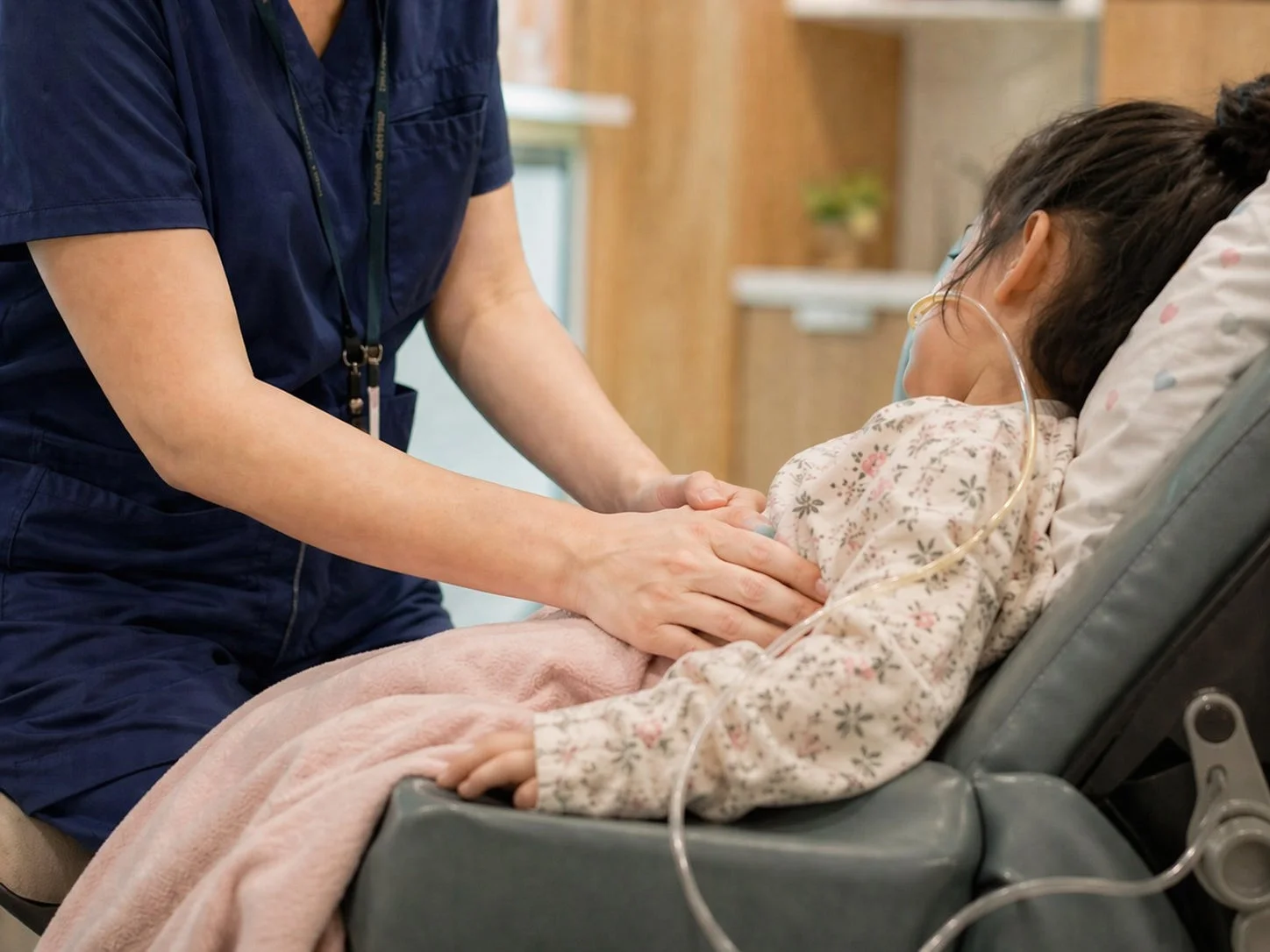 医療従事者が、鼻カニューレを付けた子供に優しく寄り添い、ケアをしている様子。子供は医療用チェアに横たわり、ピンクのブランケットに包まれている。