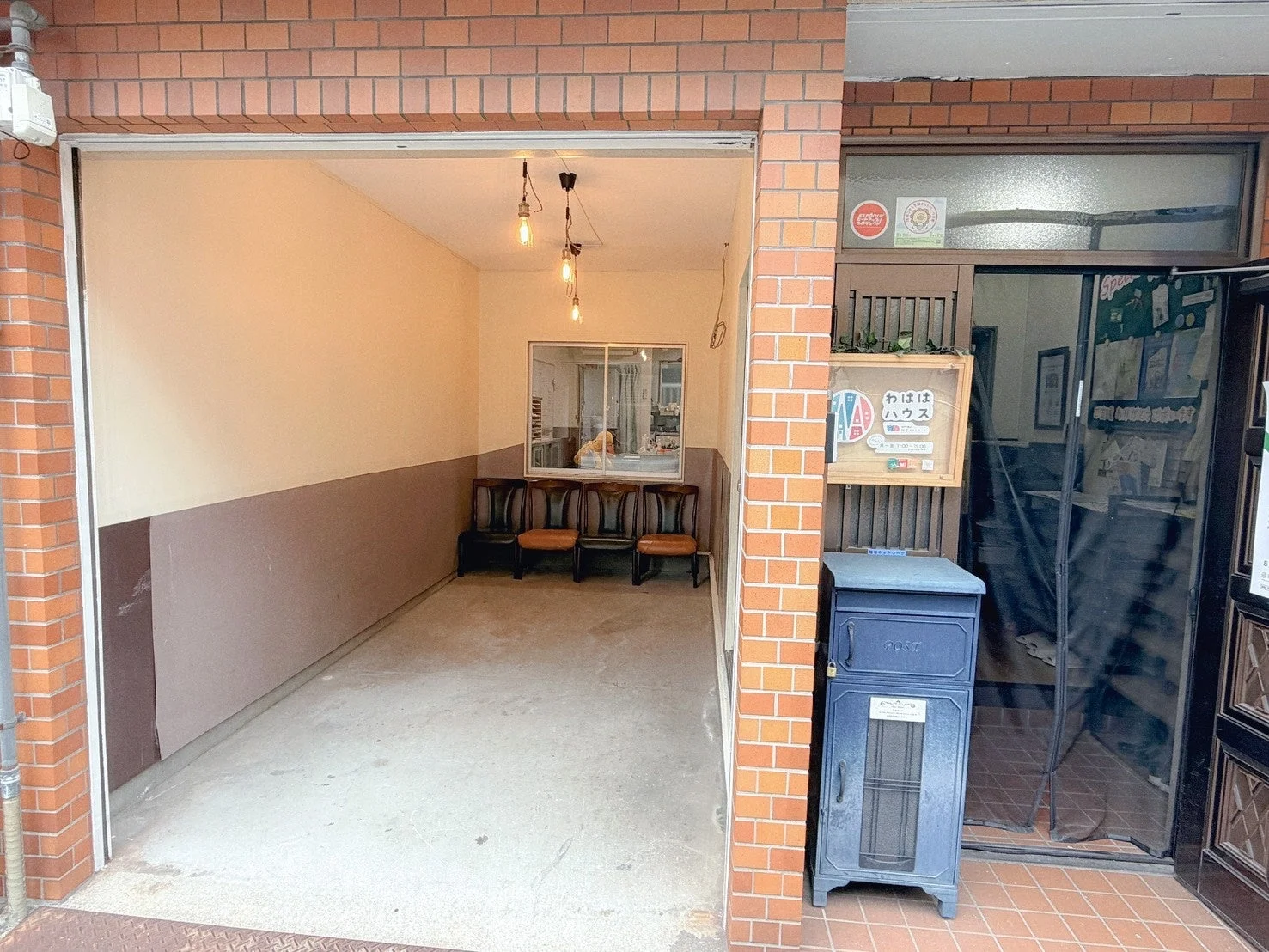 レンガ造りの建物の入口部分で、開け放たれたガレージのような空間に椅子が並び、待合室のようになっている。