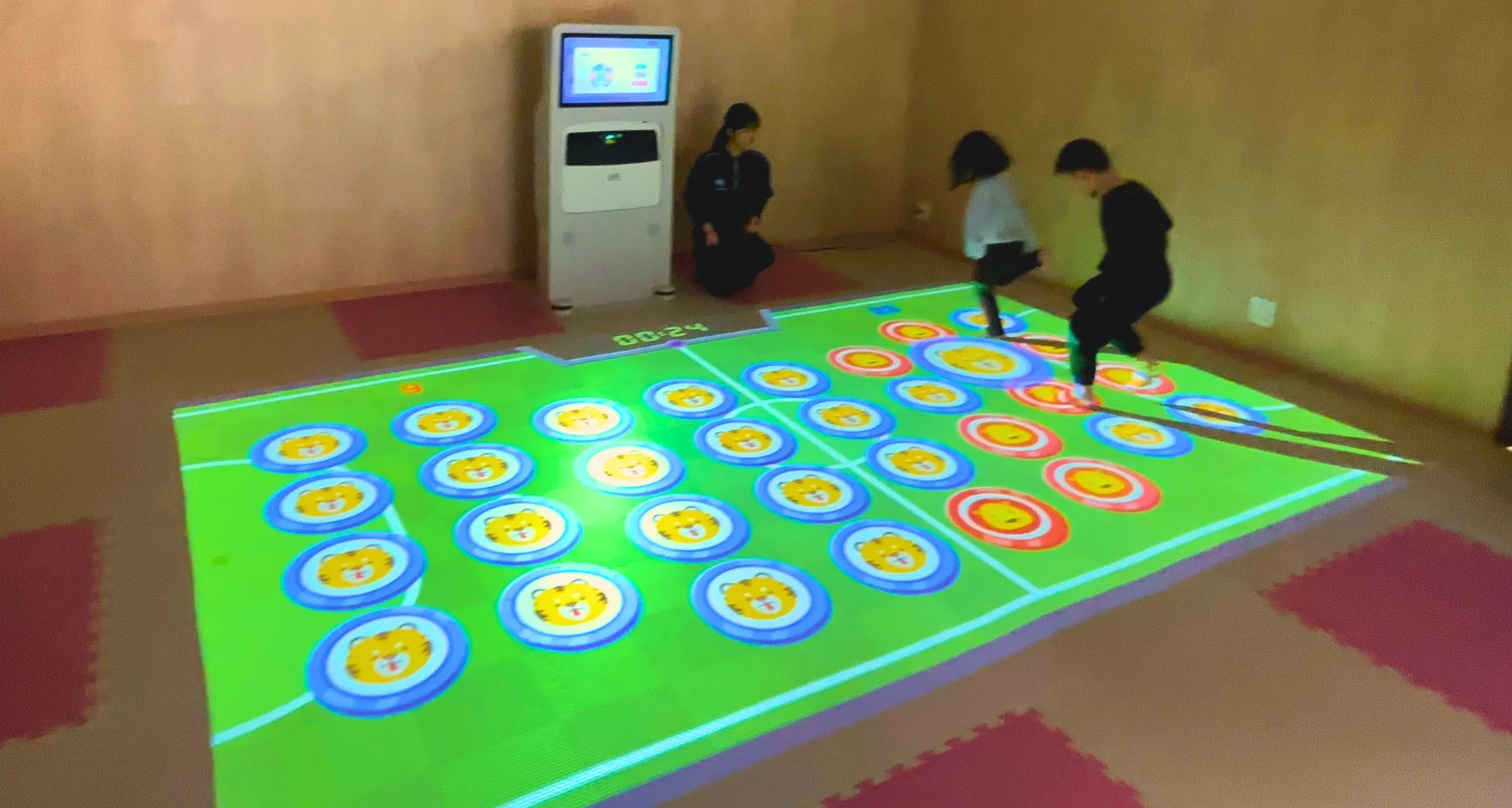 室内で子供たちが床に投影されたインタラクティブなデジタルゲームで遊んでいます。虎の顔などが描かれたカラフルな円を足で踏んで楽しんでおり、傍らには大人が見守っています。最先端の技術を使った遊び場の一コマです。
