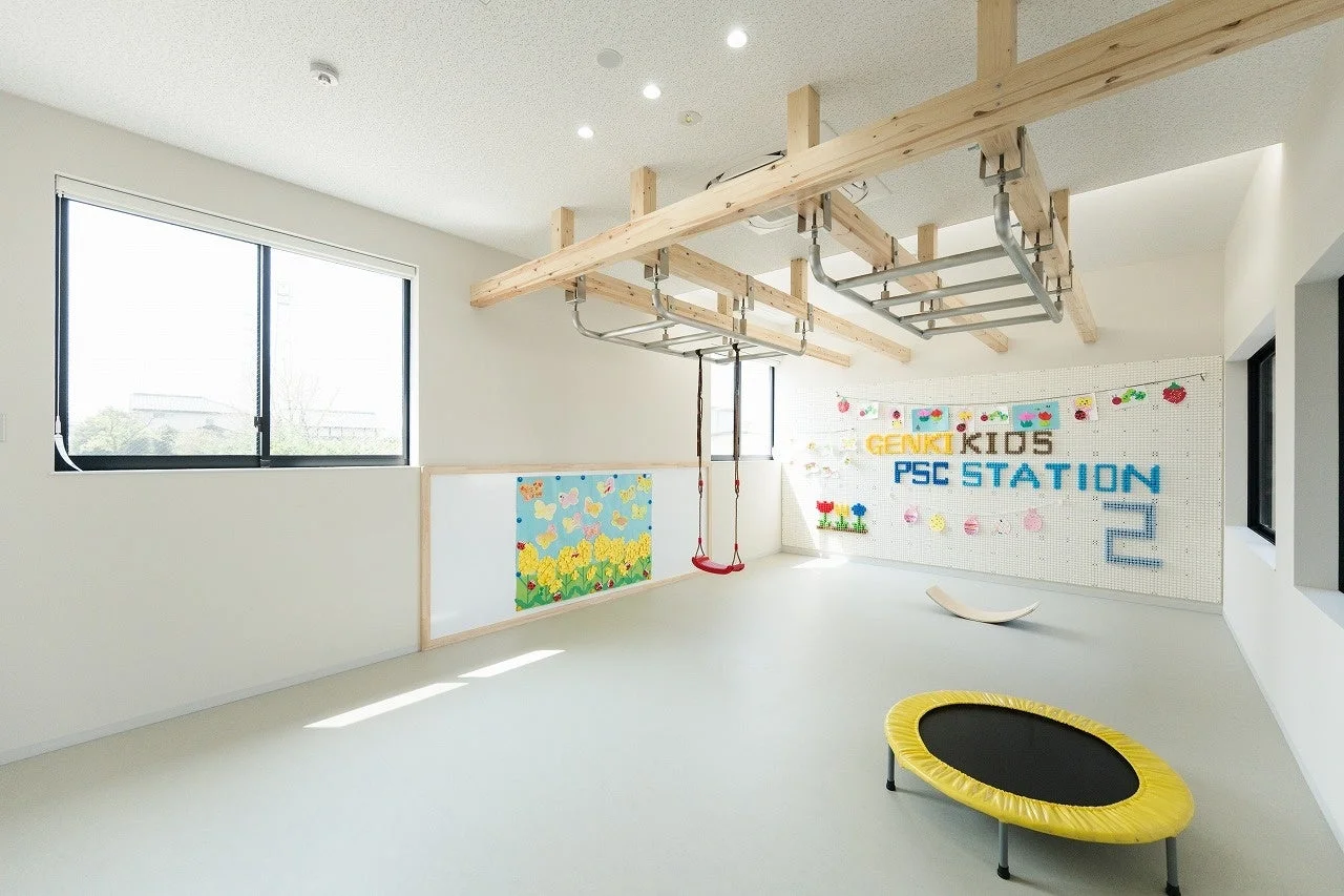 木製のうんていやブランコが設置された、明るく広々とした子供向けのプレイルームです。壁には「GENKI KIDS PSC STATION 2」の文字や可愛らしい装飾があり、床にはトランポリンやバランスボードも置かれ、子供たちが楽しく体を動かせる空間となっています。