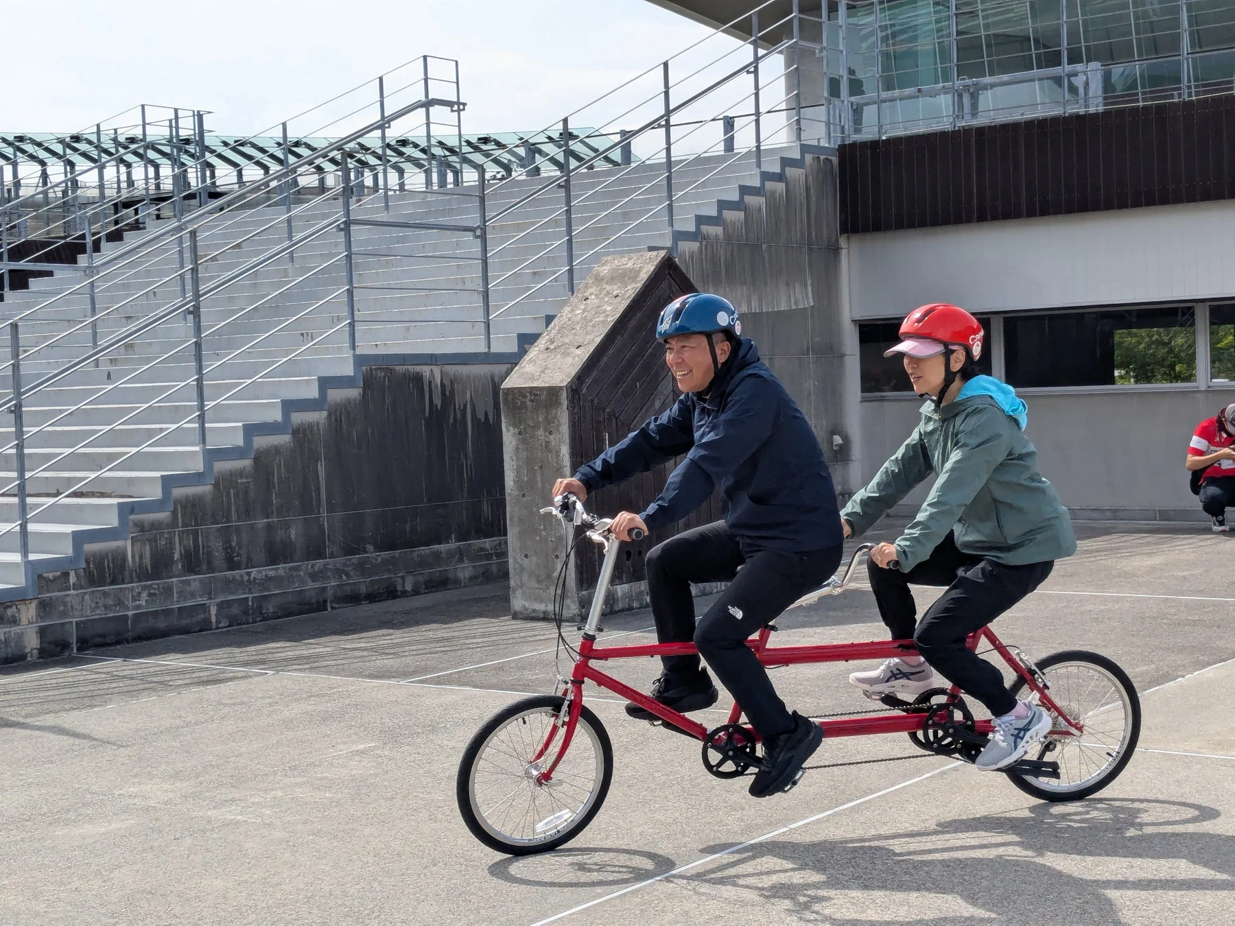 ヘルメットを着用した2人が赤いタンデム自転車に乗って屋外をサイクリングしている様子