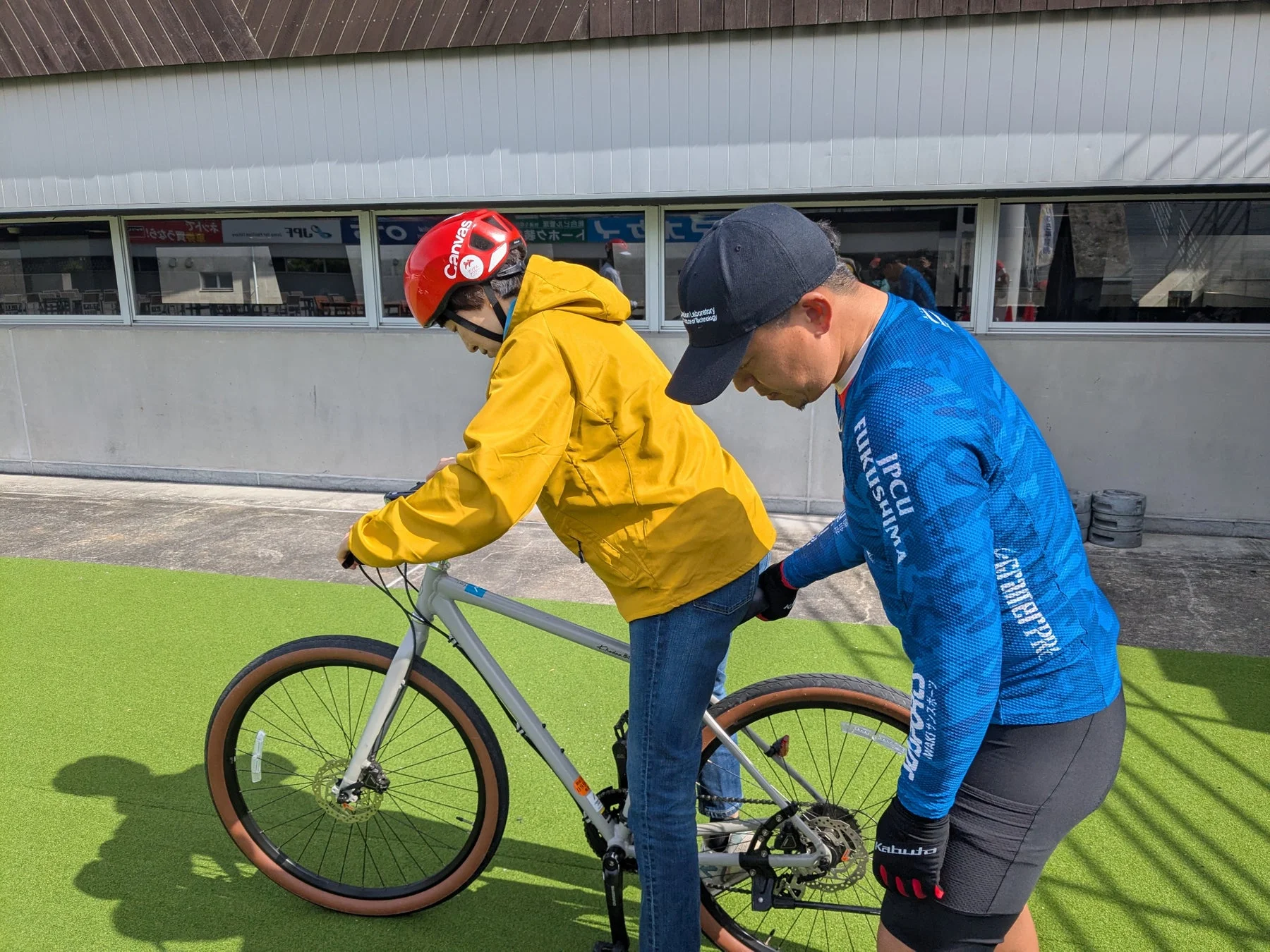 屋外の人工芝の上で、大人が子ども(または初心者)に自転車の乗り方を教えている様子