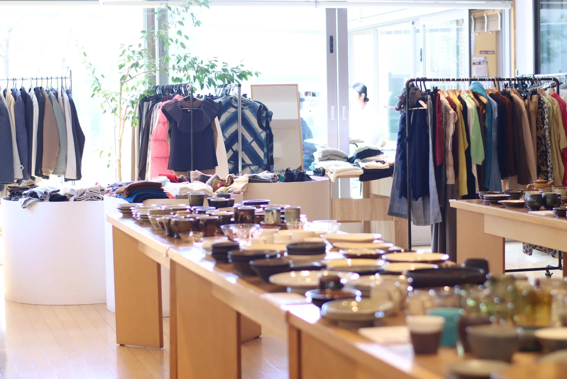明るい店内には、洋服が並んだハンガーラックと、様々なデザインの食器や陶器が陳列された木製のテーブルが複数配置されています。