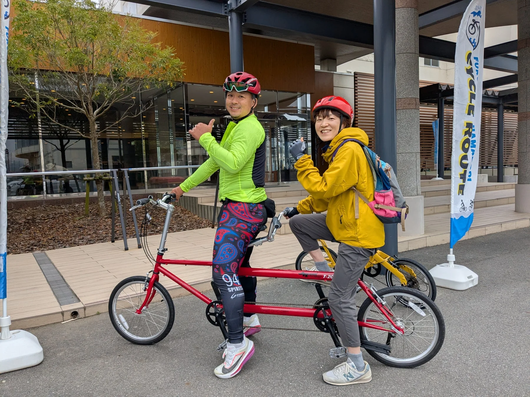 赤いタンデム自転車に乗った男女が、笑顔で親指を立ててポーズをとっています