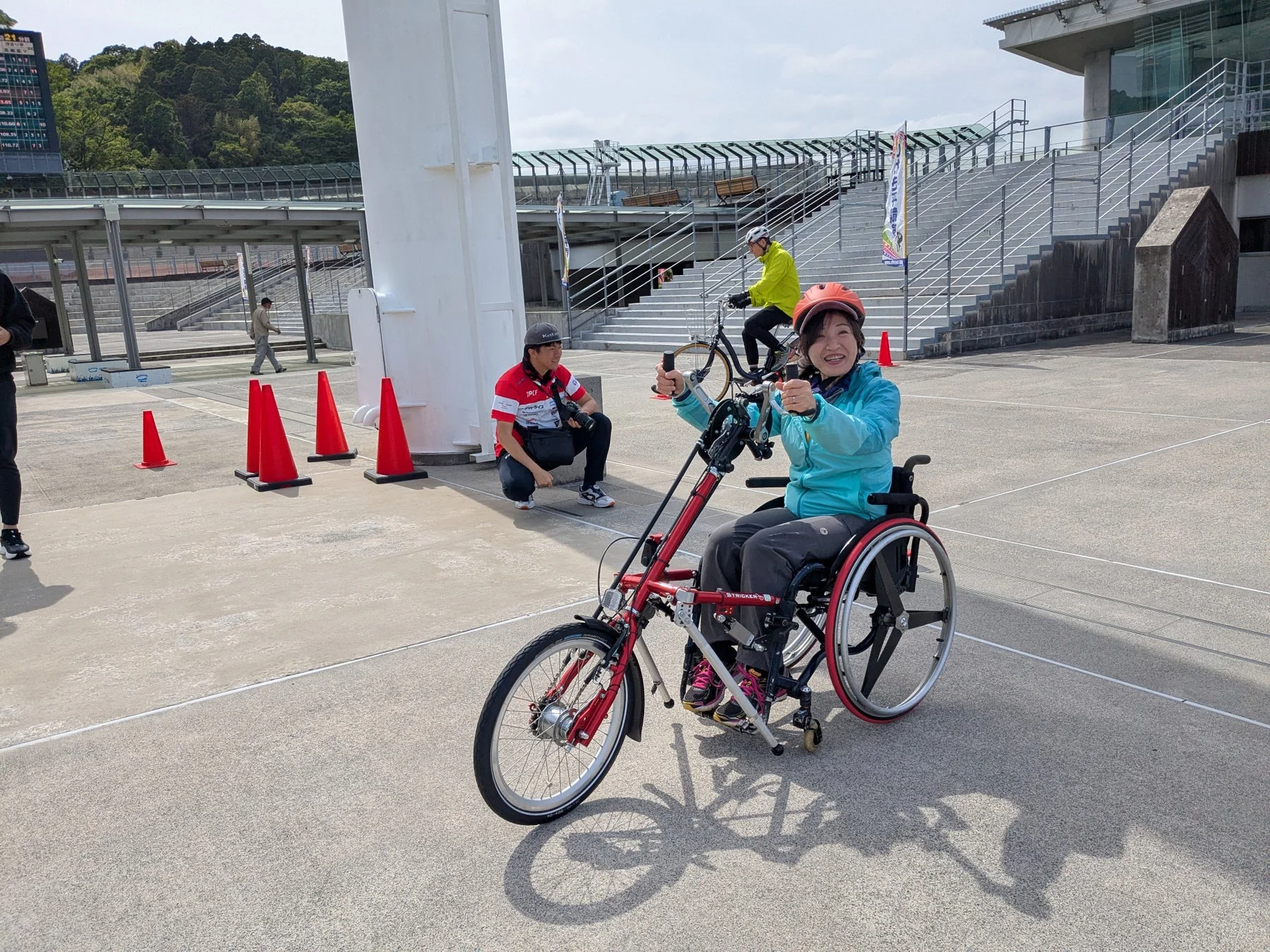 屋外の広場で、ハンドサイクルに乗った女性が笑顔でカメラを見ています