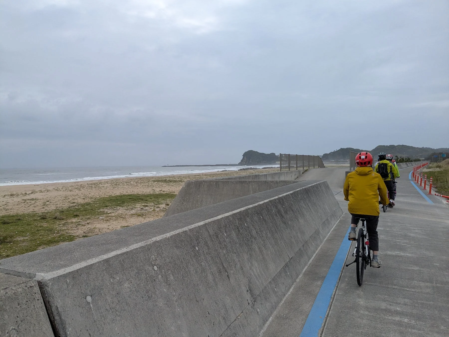 曇り空の下、数人が海沿いのサイクリングロードを自転車で走っています