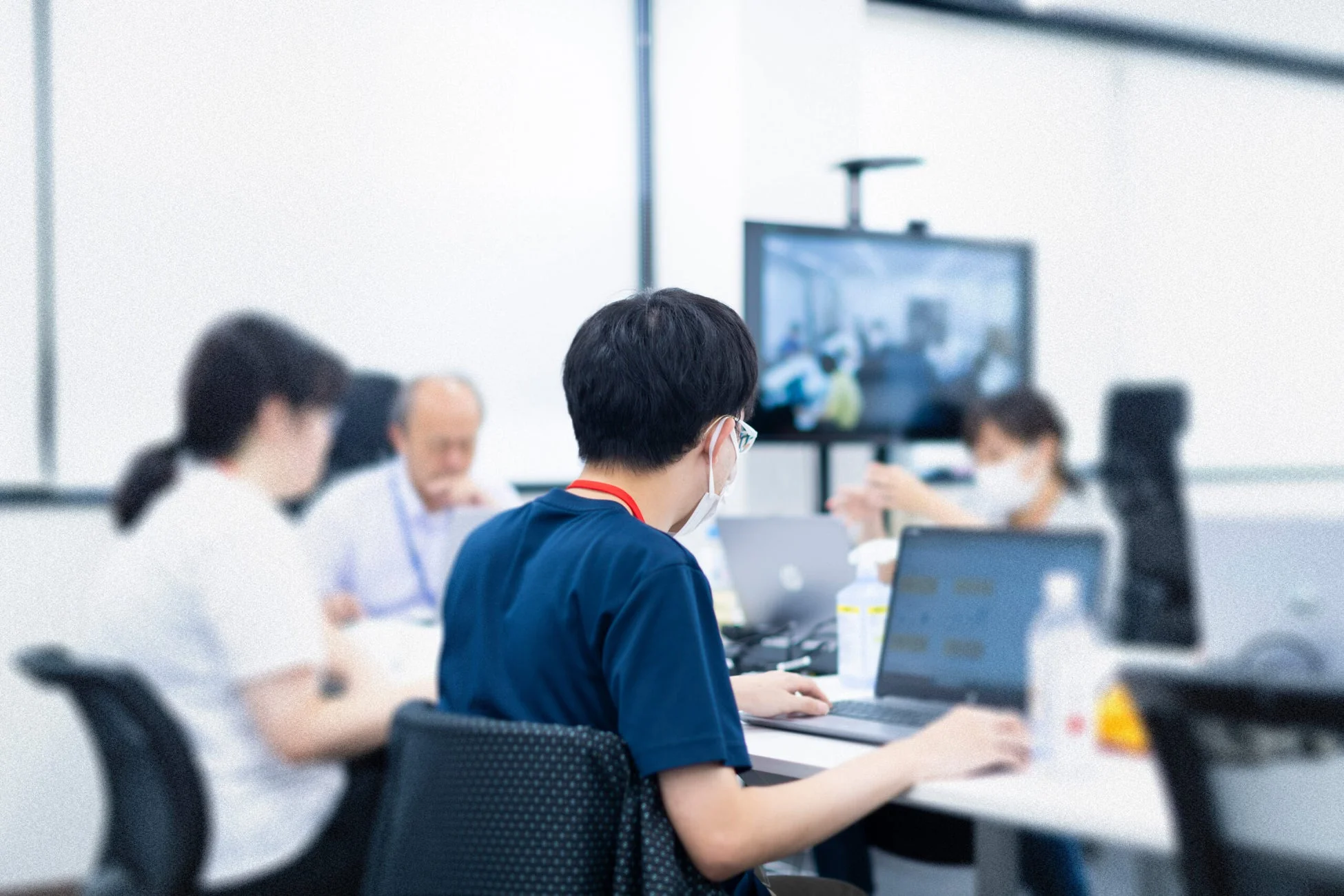 オフィスのような空間で、複数の人がノートパソコンに向かって作業している様子。手前の人物はマスクを着用し、画面の向こうにいる人とビデオ会議をしているような雰囲気も感じられます。