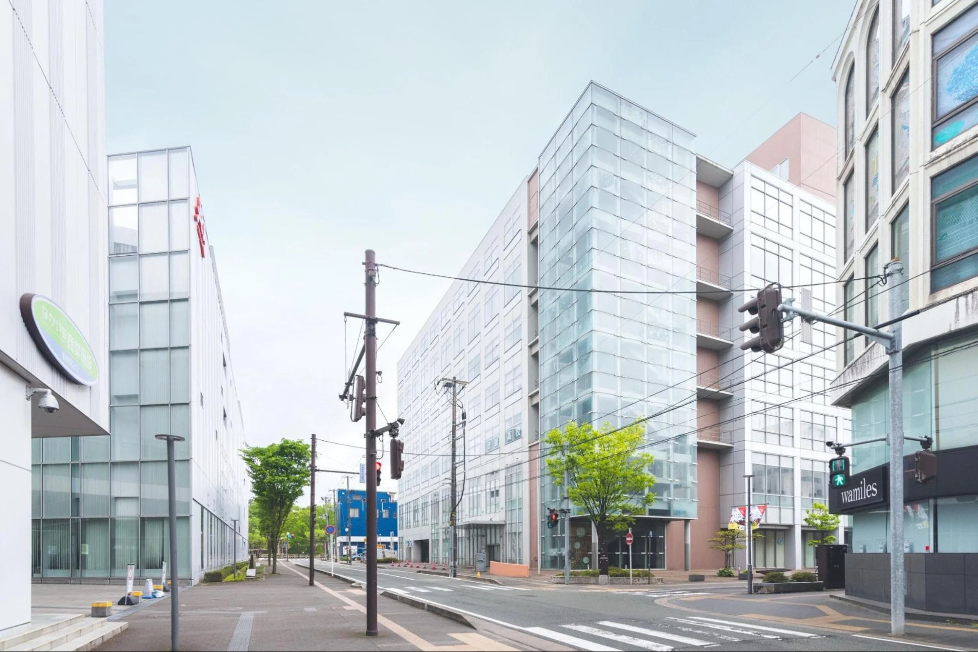 日本の都市部にあるオフィス街の風景です。近代的なビルが複数立ち並び、ガラス張りの建物が特徴的です。道路には信号機や電柱があり、清潔感のある街並みが広がっています。