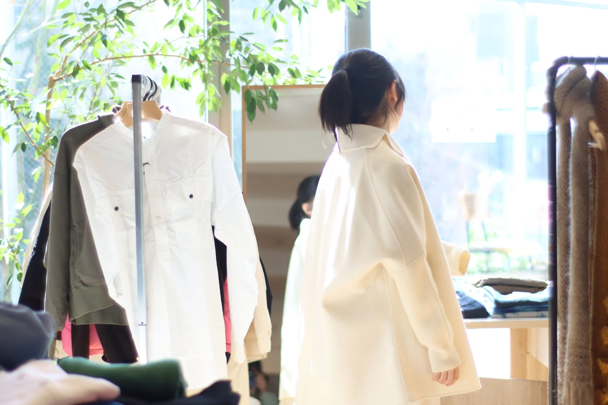自然光が差し込むアパレルショップの店内で、女性が洋服の並ぶラックの近くに立っている後ろ姿を捉えた写真。