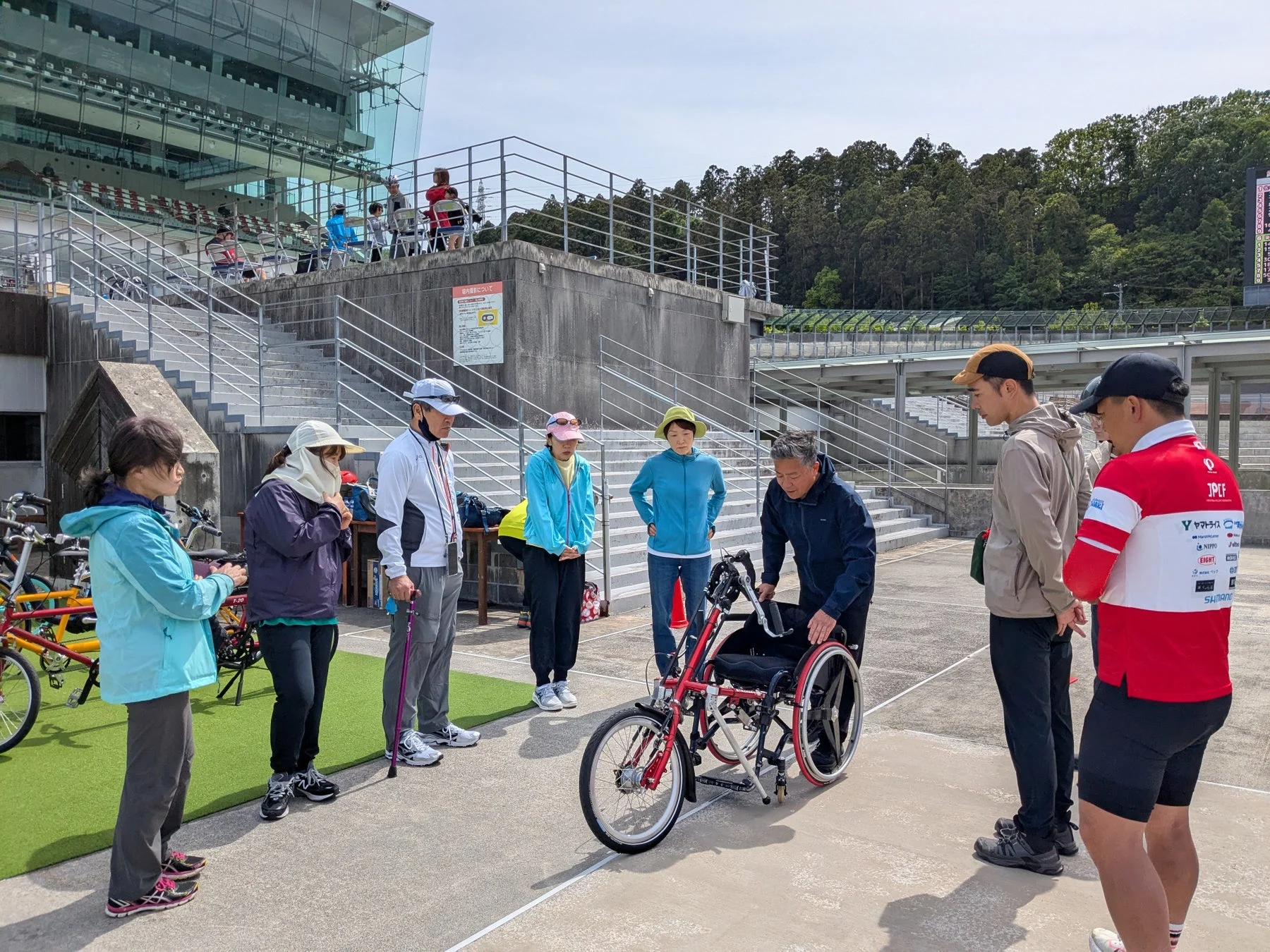 屋外のスポーツ施設で、数名の参加者が赤いハンドサイクルを囲んで説明を受けている様子