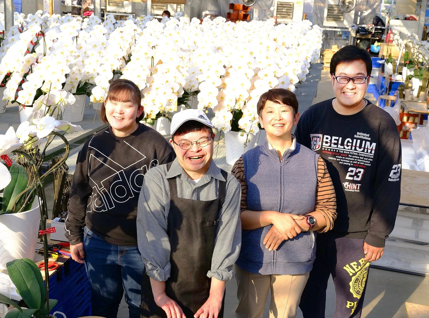 胡蝶蘭が咲き誇る温室で、4人の男女が笑顔でカメラに向かって立っています