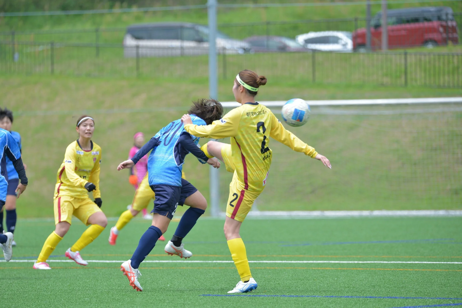 緑のサッカーフィールドで女子サッカーの試合が行われ、黄色と青色のユニフォームを着た選手たちがボールを蹴り合っている様子