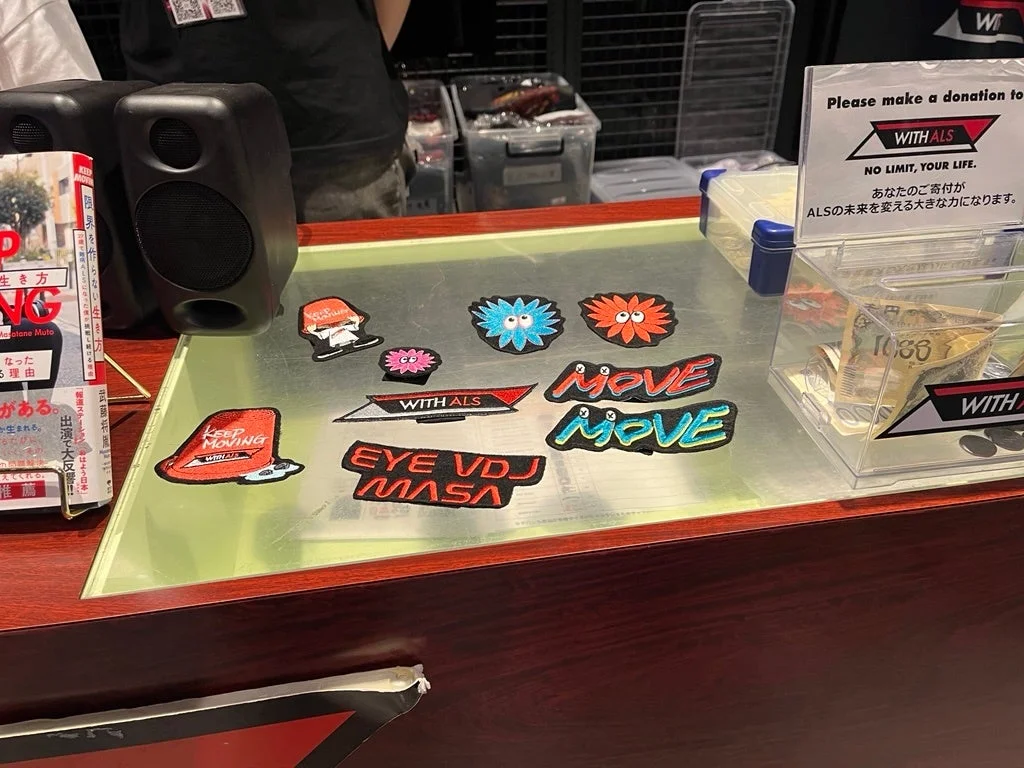 ALS支援と啓発のための募金活動ブース。募金箱には「WITH ALS」のメッセージと日本円が、テーブルには「KEEP MOVING」などのワッペン、書籍、スピーカーが展示されている。