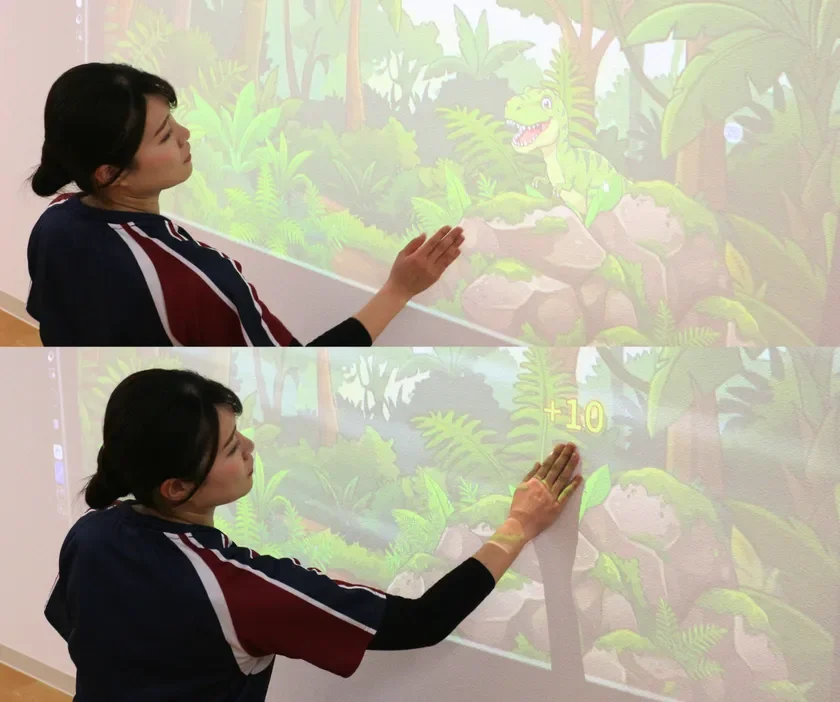 女性が壁に投影されたジャングルの恐竜の画像とインタラクトしている様子。手をかざすと「+10」と表示され、インタラクティブなゲームや学習コンテンツを示唆しています。