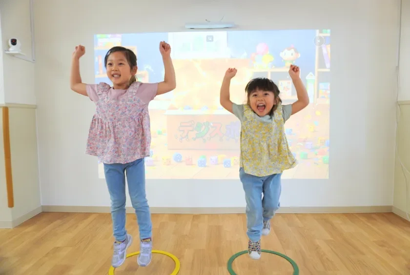 2人の女の子が、室内のプロジェクターに映し出された映像に合わせて、フープの中で楽しそうにジャンプしている様子。満面の笑みを浮かべ、元気に体を動かしている場面です。