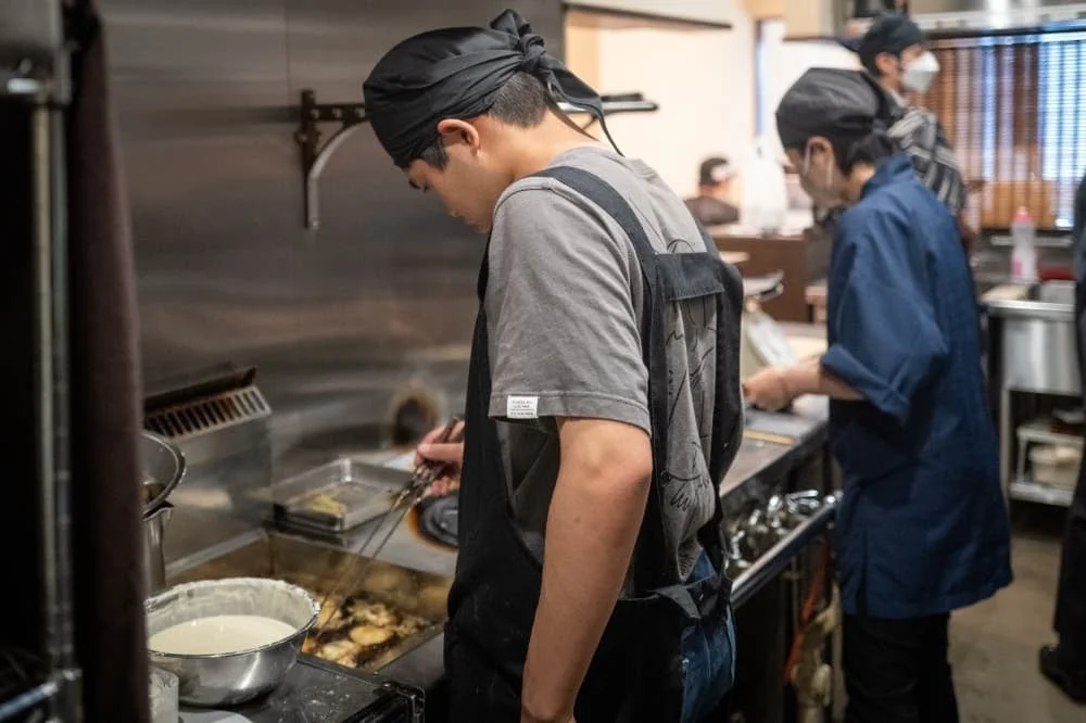 飲食店らしき厨房で、黒いバンダナを巻いた料理人が揚げ物をしており、その隣には別のスタッフも作業している様子が写っています。