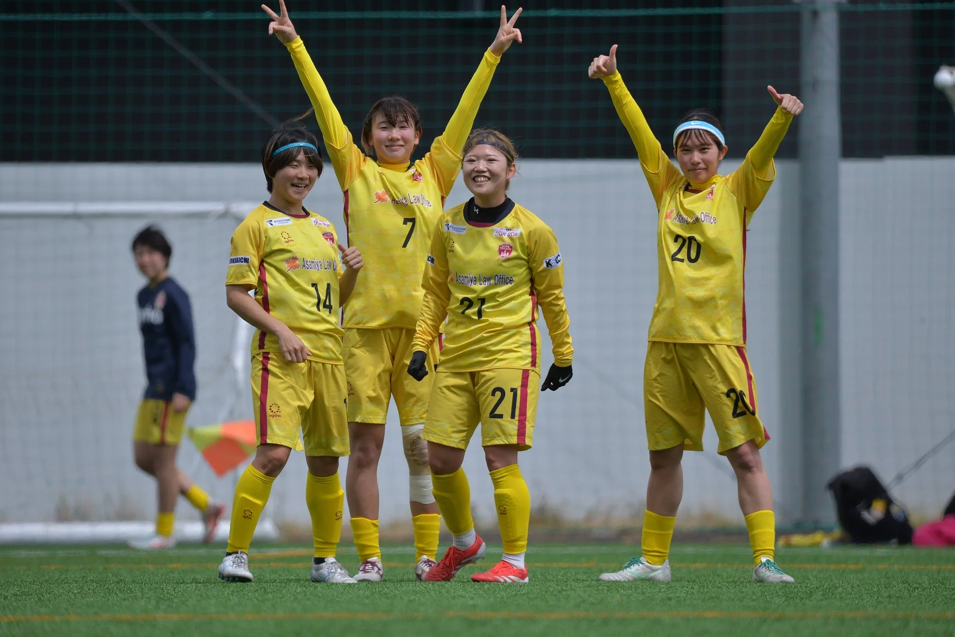 黄色いユニフォームを着た4人の女子サッカー選手が、緑のグラウンドで笑顔でポーズをとっている様子