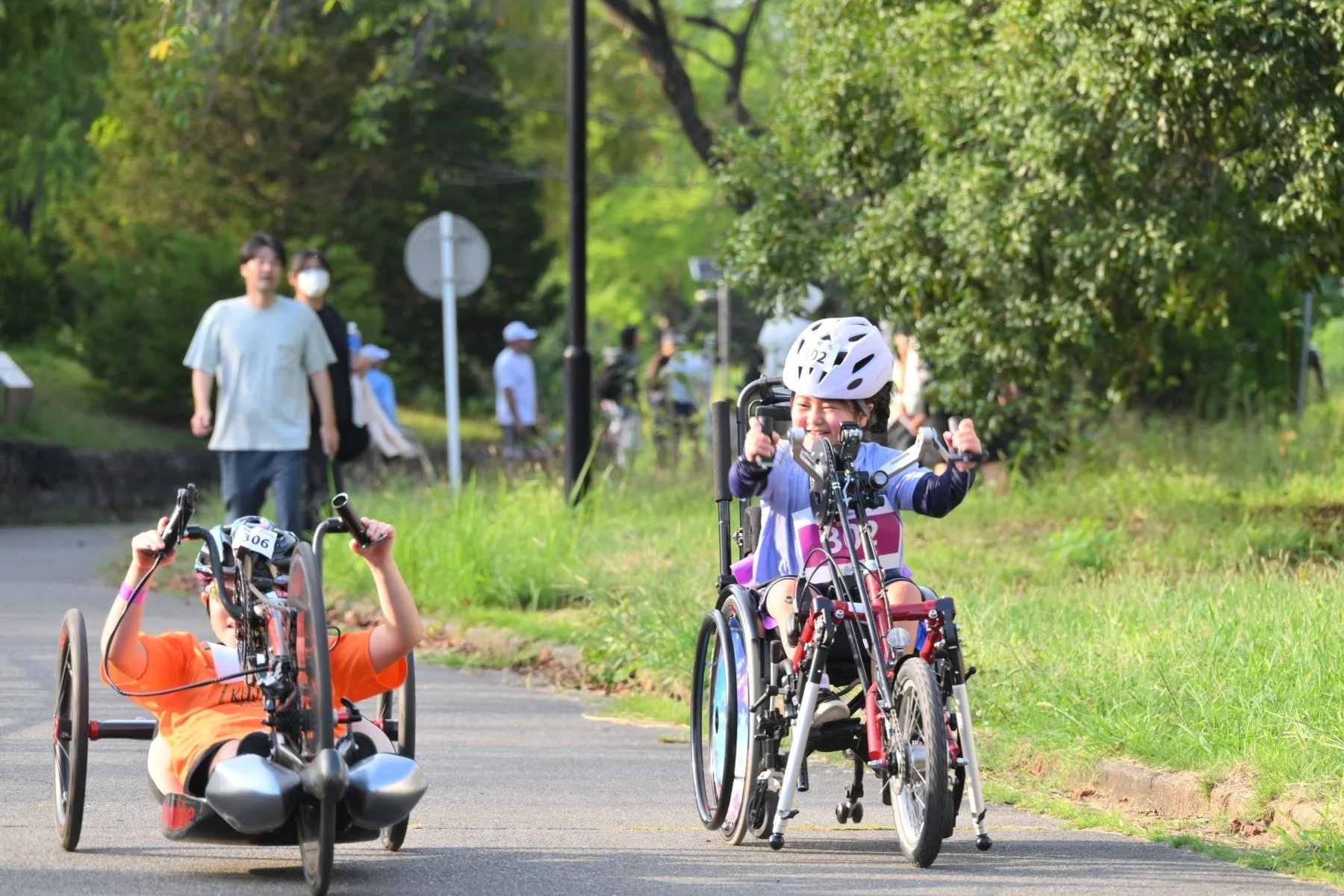 公園の舗装された道で、子供がハンドサイクルに乗って楽しそうに走っている様子