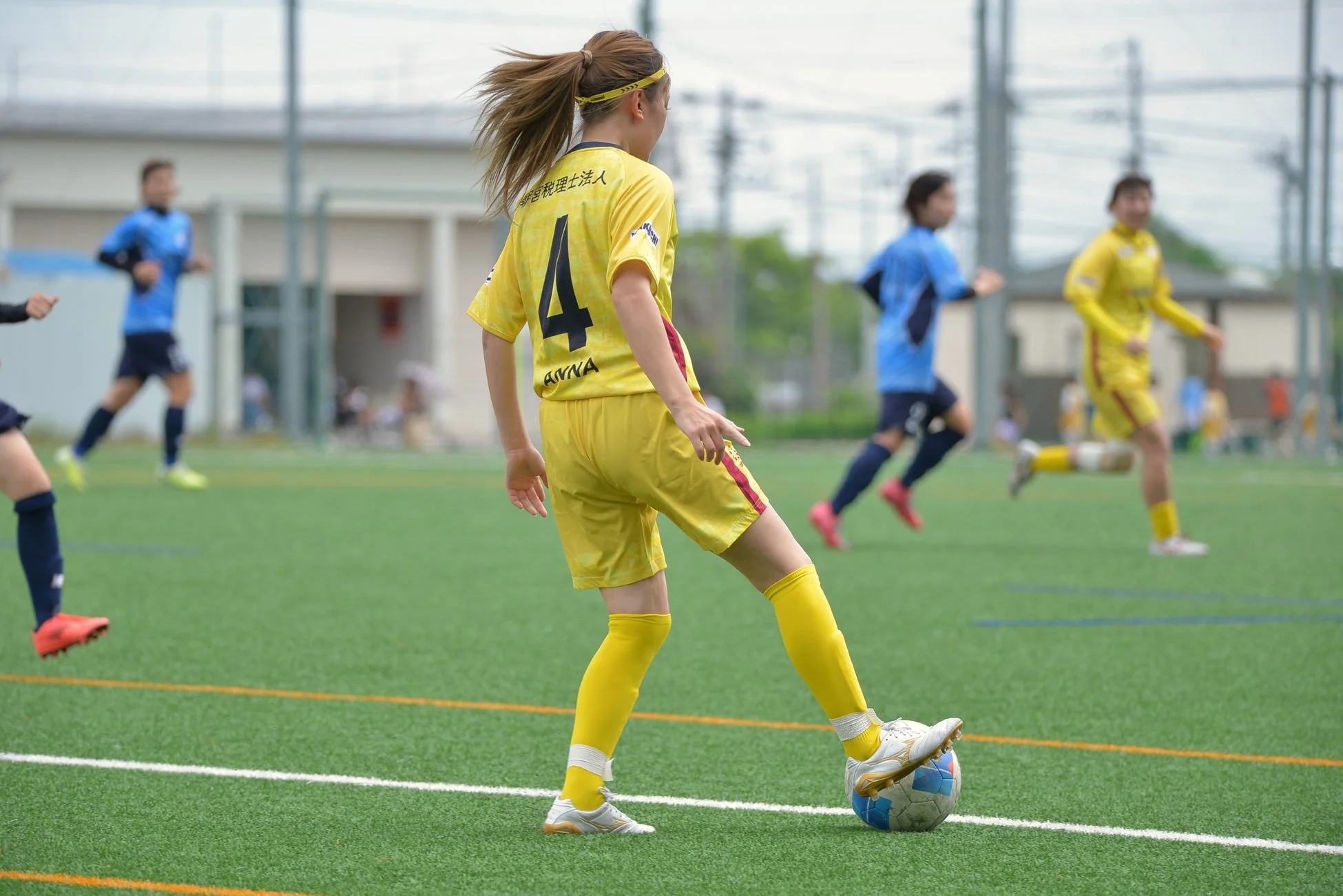 人工芝のサッカーグラウンドで、黄色いユニフォームを着た女性選手がボールをドリブルしている様子