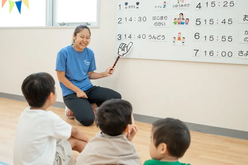 女性の先生が笑顔で、床に座っている3人の子供たちにホワイトボードに書かれた一日のスケジュールを説明しています。指差し棒を使って熱心に話しており、子供たちは集中して耳を傾けています。