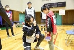 体育館のような場所で、大人がマスクを着用ししゃがんで、赤いベストを着た子どもに何かを指導している場面。