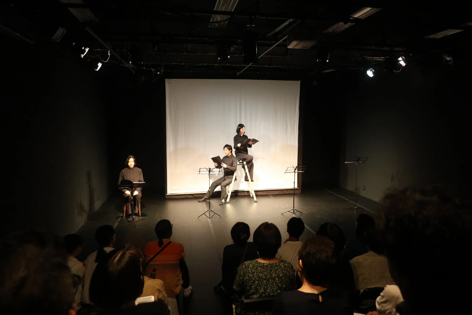 暗い舞台で朗読パフォーマンスを行う3人の演者