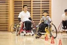 体育館で車椅子に乗った大人と子供が、オレンジ色のコーンを使って車椅子操作の練習をしている様子。