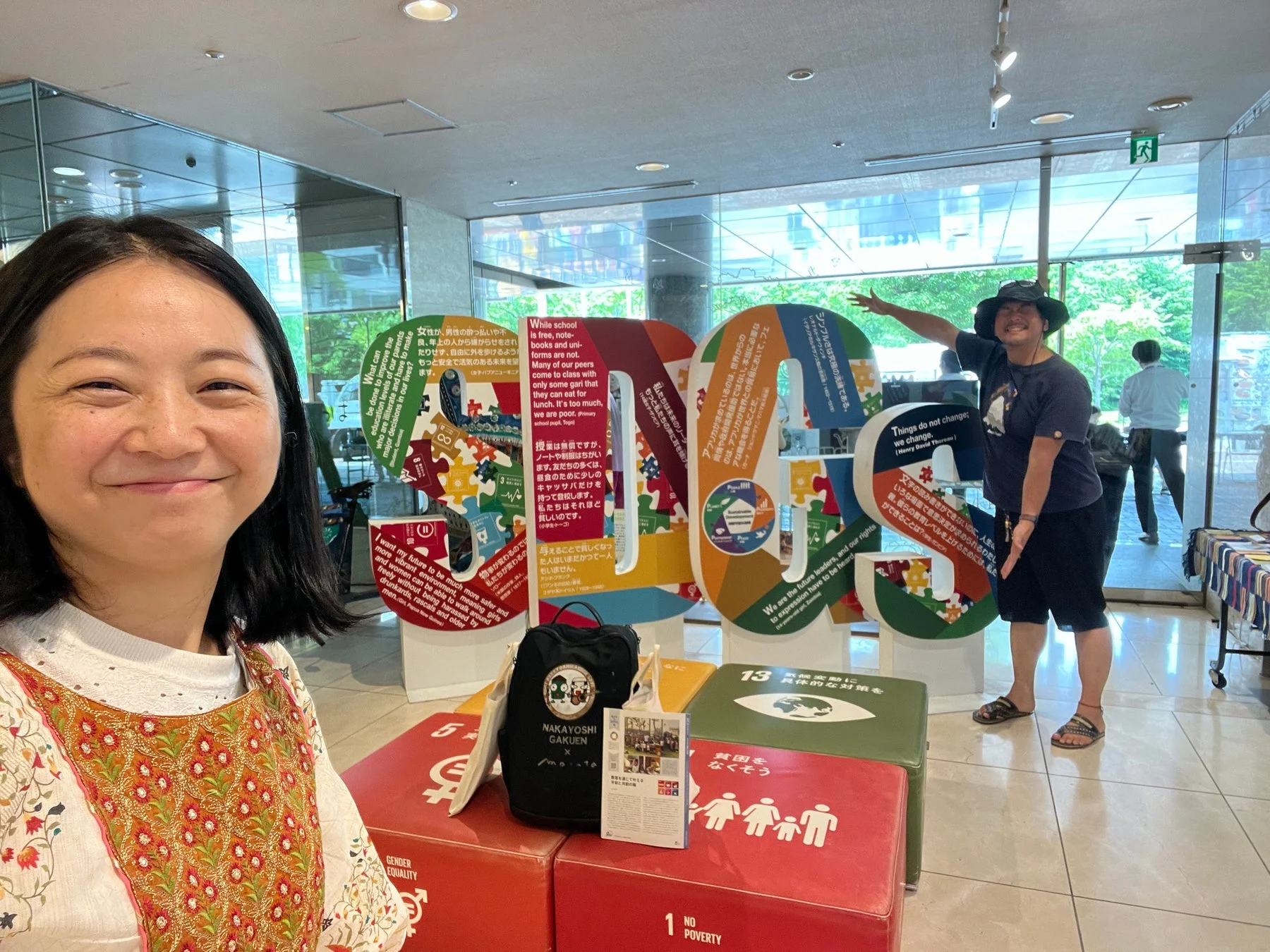 SDGsに関する展示イベントで、女性2人が笑顔でポーズをとっています。カラフルなロゴや、貧困、教育、ジェンダー平等、気候変動など、世界各地からのメッセージが書かれたパネルが設置されています。国際協力や社会貢献への意識を高めるための催しです。